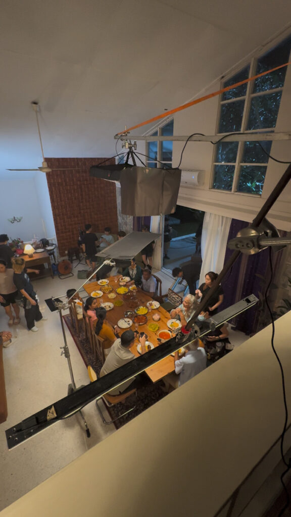 Wide view from a mezzanine of a long family dinner table with lights and crew around it