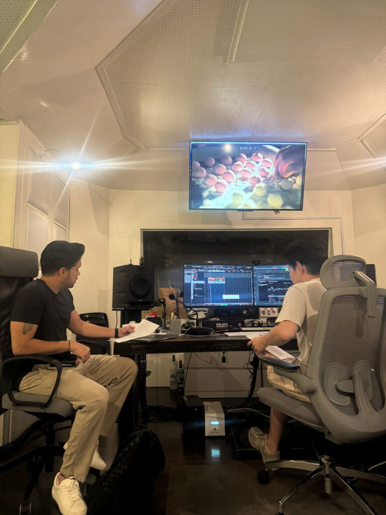 Two people in a sound studio reading scripts, with the film on a screen above them